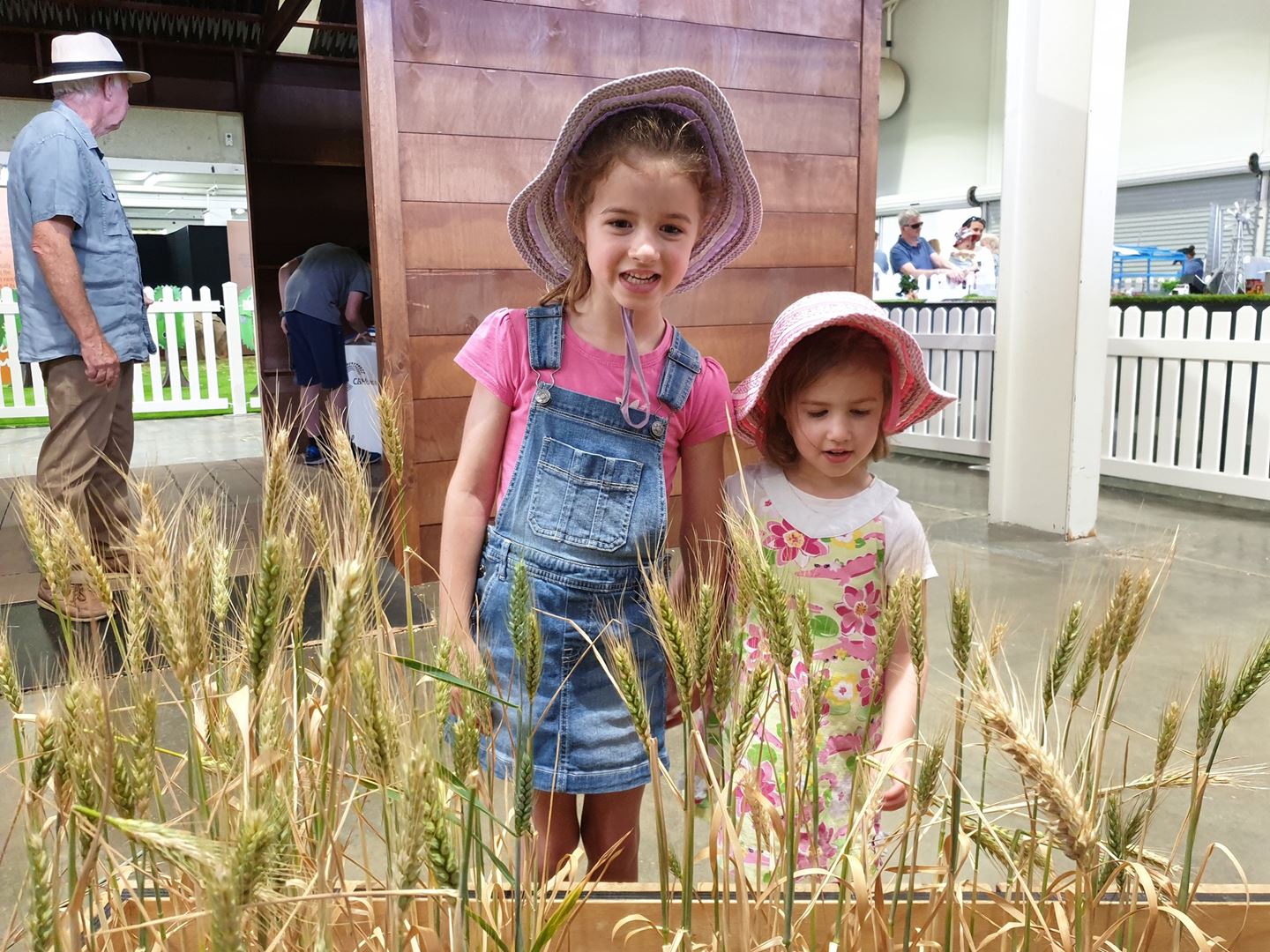 Two young girls wearing floral hats stand behind a planter box of wheat smiling at the camera