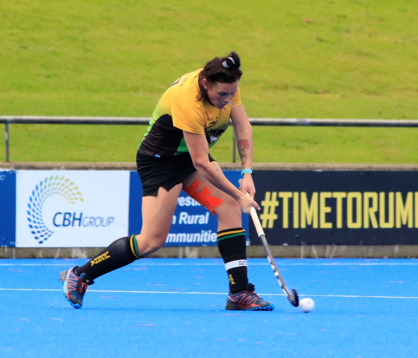 A girl wearing a yellow and green uniform swings a hockey stick