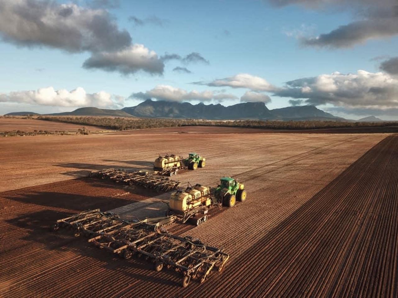 Two green tractors pulling two seeders work side by side in a reddish brown paddock