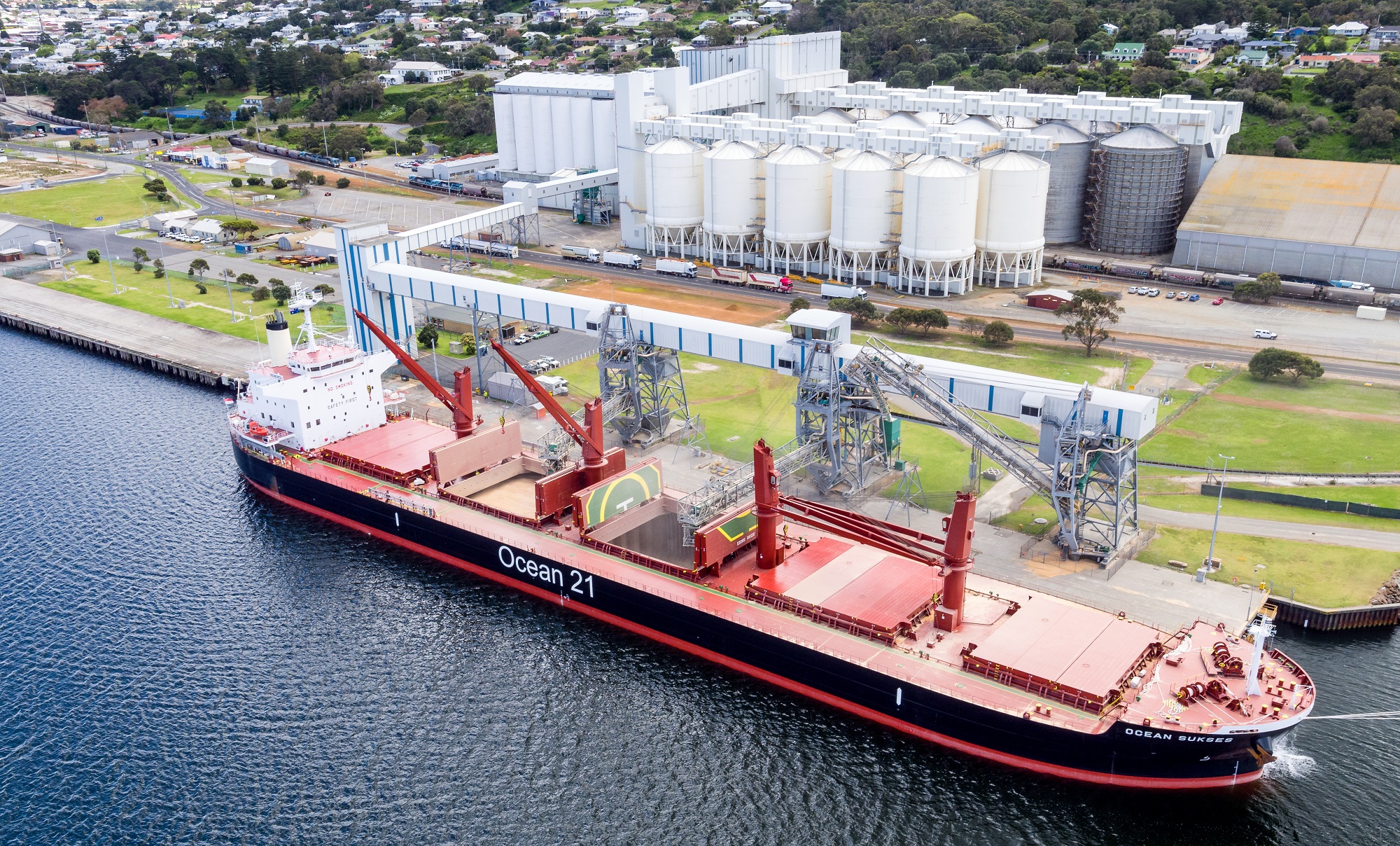A red ship is anchored at a wharf with white grain silos in the background