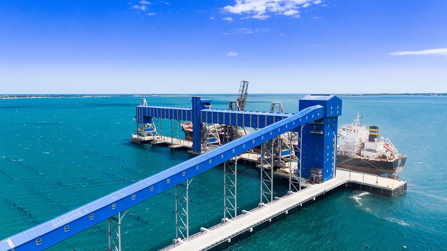 A large ship is anchored next to a long blue wharf with clear blue skies and turquoise ocean in the background