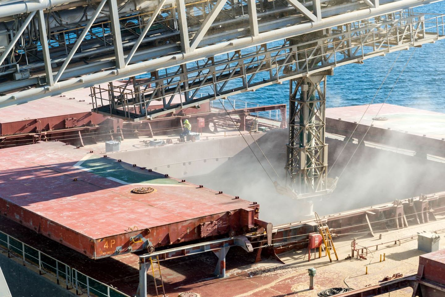 A shiploading structure discharges grain into the open hatch of a ship, grain dust is flying everywhere
