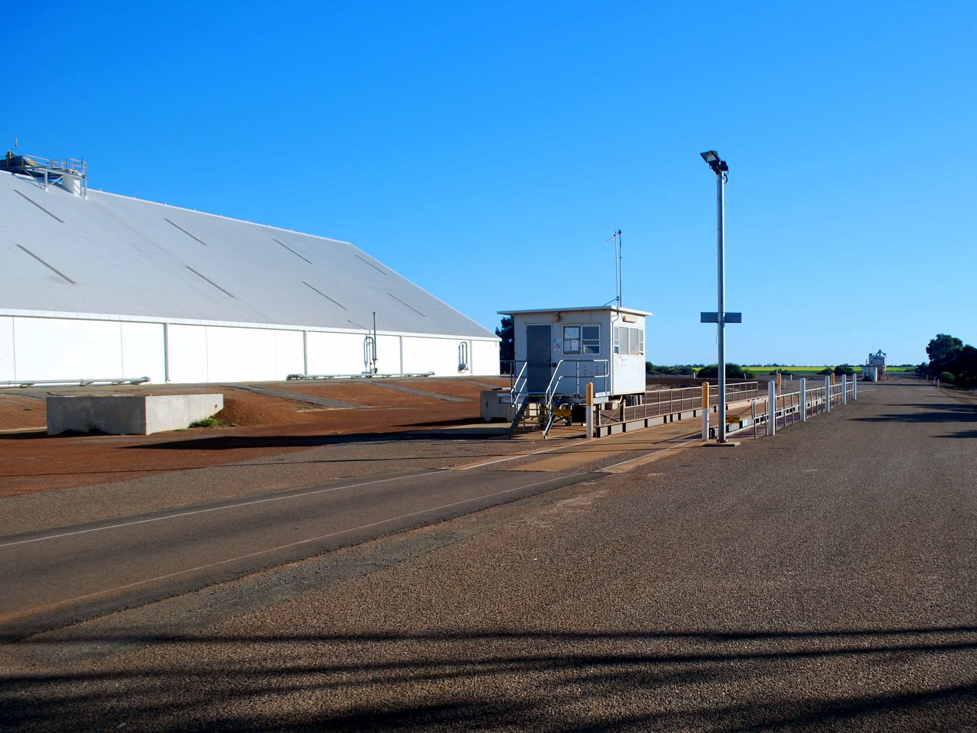 Image of CBH Yuna sample hut with blue sky and horizontal storage shed in the background, bitumen road in the foreground