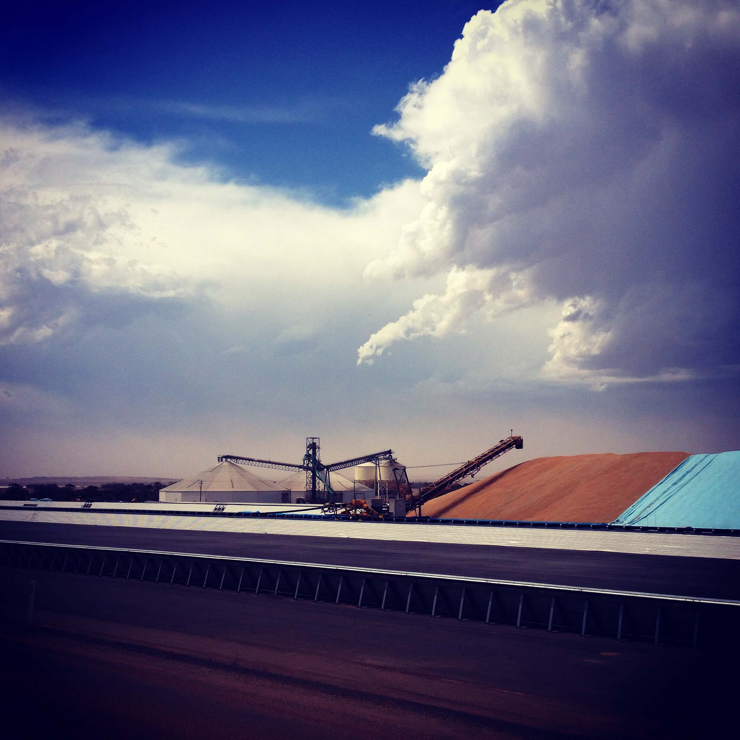 Panoramic shot of grain being loaded in to an open bulkhead with large puffy white clouds overhead. The lower third is the asphalt of the road.