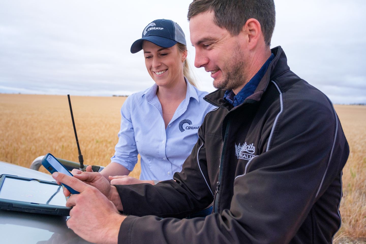 A female CBH Business Relationship Manager helps a grower contract online using his mobile phone at the front of a car in a paddock