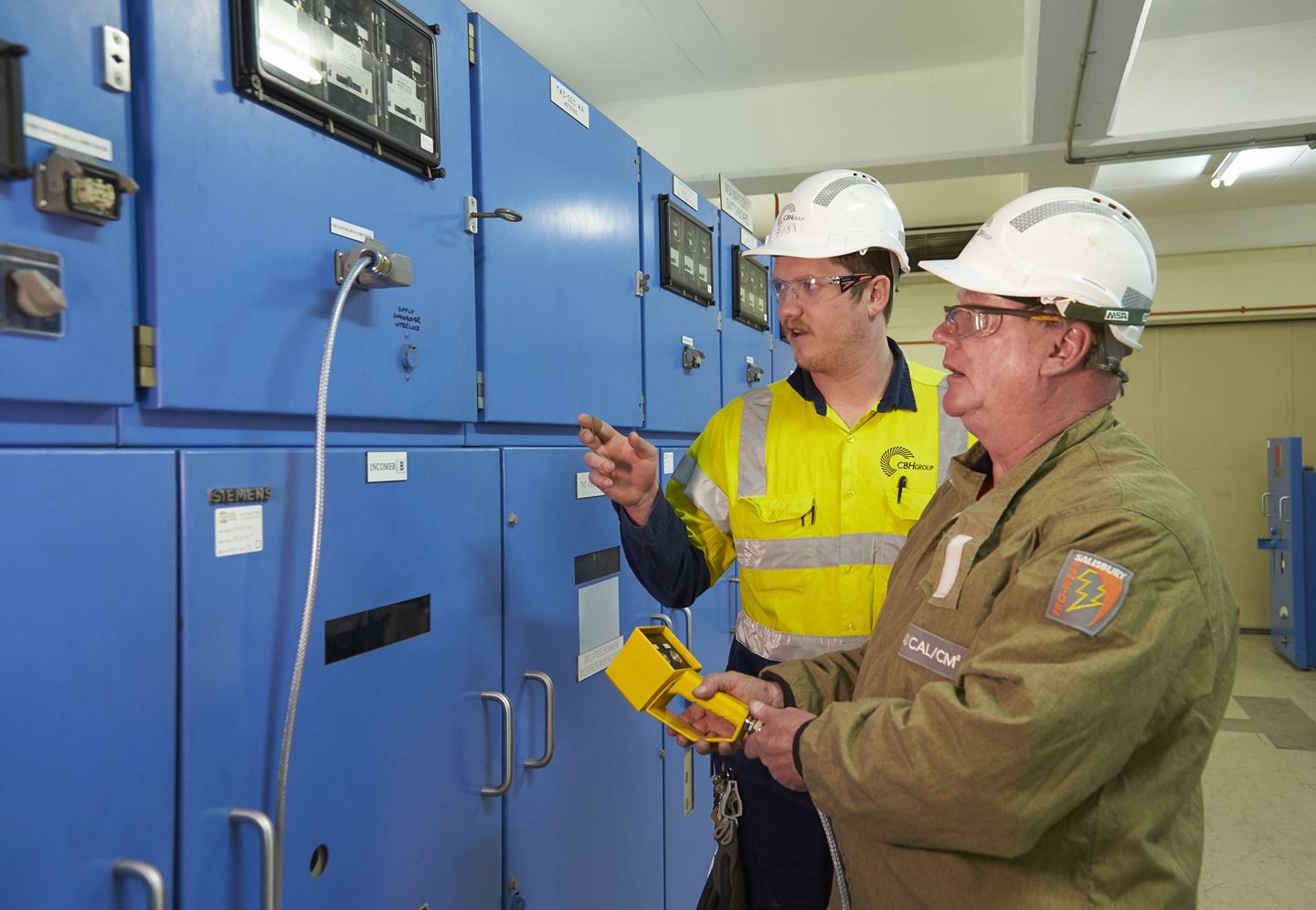 Two people in hard hats inspecting a switch board