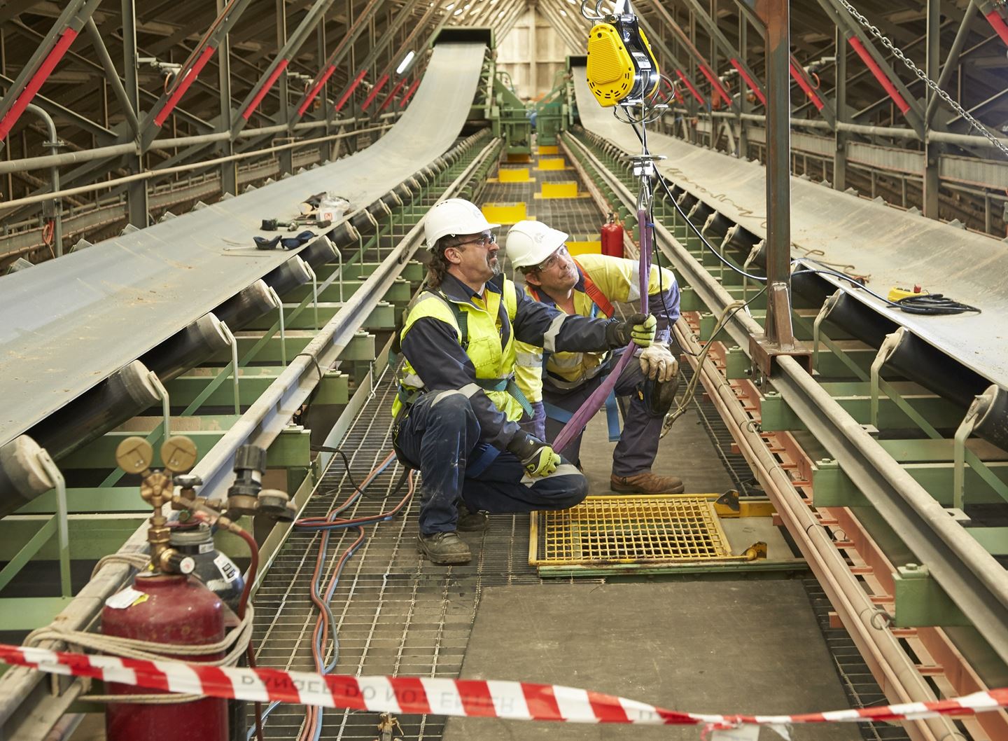 Two men in work clothing inspect a strap between two conveyor builts