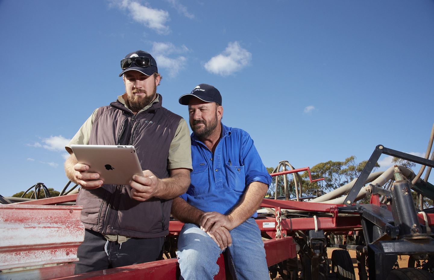 Two men wearing caps sit on a red tractor while looking at an ipad together