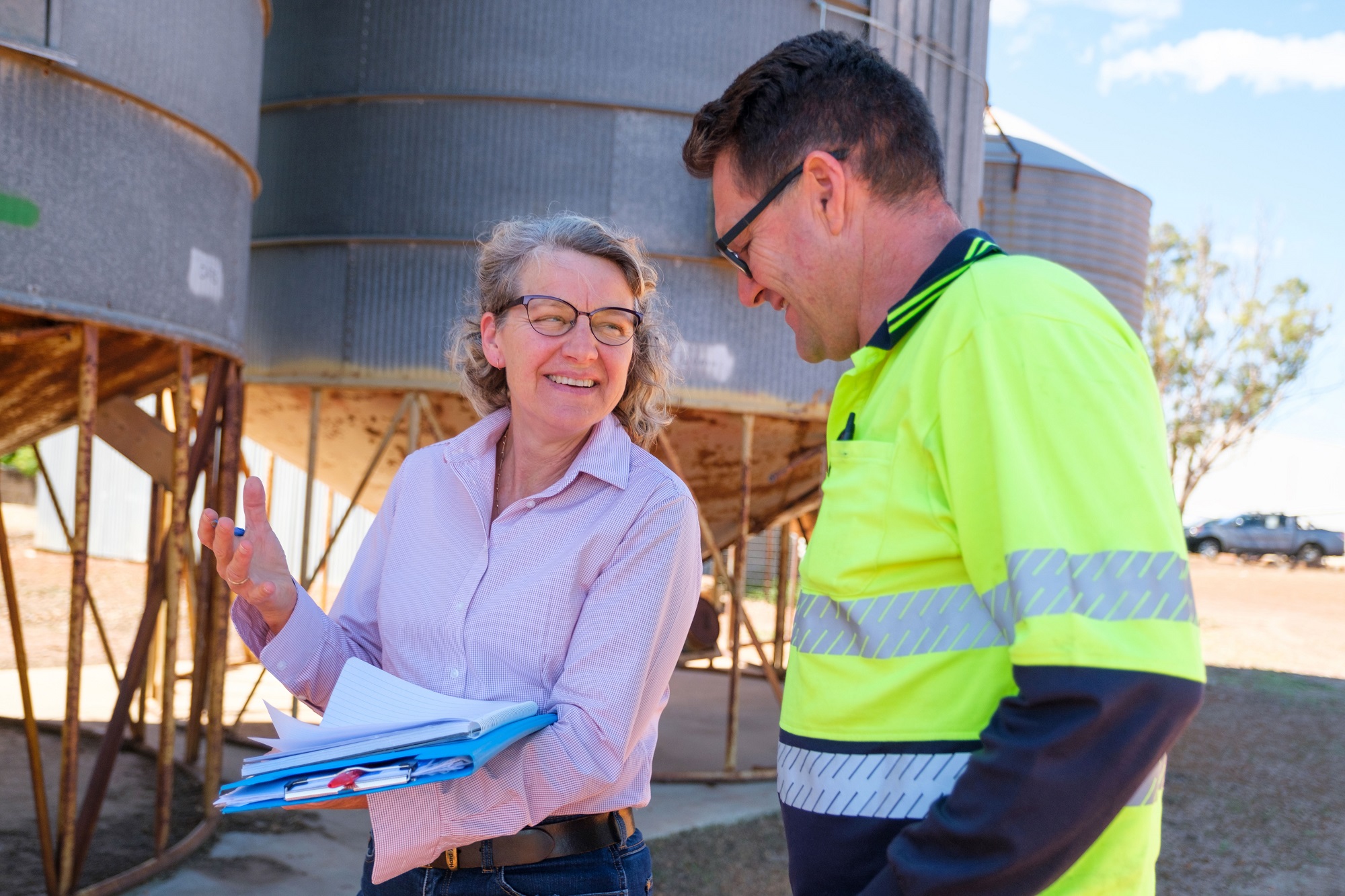 A woman holding a clipboard smiles at a man wearing a hi vis yellow shirt during his ISCC audit