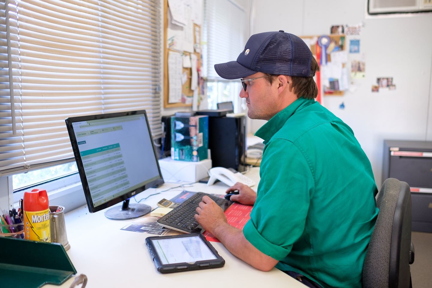 A man in a green shirt and navy cap sits at a computer